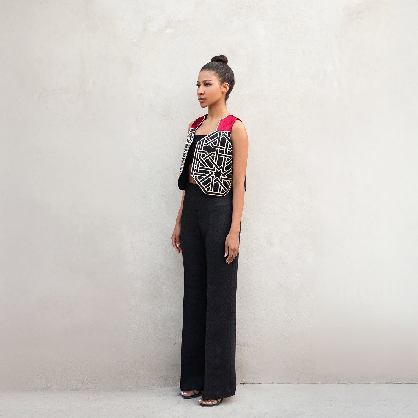 A side profile of the same outfit, showing the sleeveless velvet vest with crimson shoulders, golden geometric design, and black trousers.